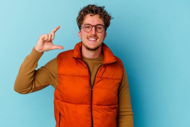 Mavi arka planda gözlük takan genç beyaz adam işaret parmaklarıyla küçük bir şey tutuyor, gülümsüyor ve kendinden emin..