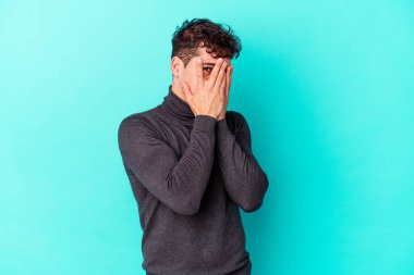Kafkasyalı genç adam mavi arka planda izole edilmiş. Korkmuş ve gergin parmaklarıyla göz kırp..