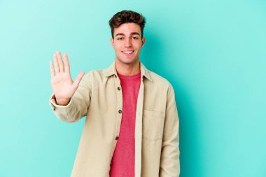 Mavi arka planda izole edilmiş genç beyaz adam gülümseyerek beş numarayı parmaklarıyla gösteriyor..