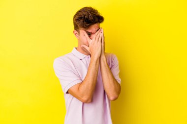 Sarı arka planda izole edilmiş genç beyaz adam parmaklarının arasından korkmuş ve gergin bir şekilde göz kırp..