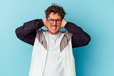 Gözlüklü genç beyaz adam mavi arka planda izole edilmiş kulakları elleriyle kapatıyor ve çok fazla ses duymamaya çalışıyor..