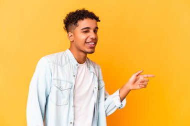 Genç Afro-Amerikan kıvırcık adamı, mor üzerine izole edilmiş, mutlu bir şekilde işaret parmağıyla işaret ederek gülümsüyor..