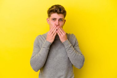 Sarı arka planda izole edilmiş genç beyaz adam elleriyle ağzını kapatırken şok oldu..