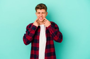 Mavi arka planda izole edilmiş genç beyaz adam gülümsüyor, parmaklarını ağıza doğrultuyor..