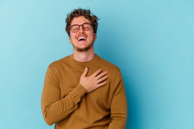 Mavi arka planda gözlüklü genç beyaz adam mutlu bir şekilde gülüyor ve ellerini karnının üstünde tutarak eğleniyor..
