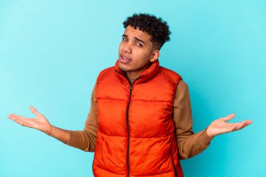 Genç Afro-Amerikan kıvırcık adamı sorgulama jesti için mavi üzerine izole edilmiş şüpheci ve omuz silkiyor..