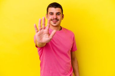 Sarı arka planda izole edilmiş genç beyaz adam gülümseyerek beş numarayı parmaklarıyla gösteriyor..