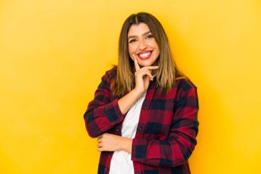 Sarı arka planda izole edilmiş genç Hintli kadın gülümsüyor mutlu ve kendinden emin, elleriyle çenesine dokunuyor..