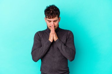 Mavi arka planda izole edilmiş genç beyaz bir adam el ele tutuşup, ağzının kenarında dua ediyor, kendinden emin..