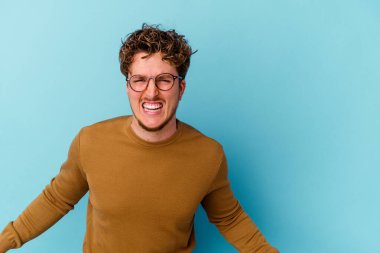 Mavi arka planda izole edilmiş gözlüklü genç beyaz adam çok kızgın, öfkeli, hayal kırıklığına uğramış.