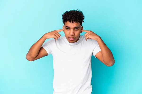 Young african american curly man isolated on blue covering ears with fingers, stressed and desperate by a loudly ambient.