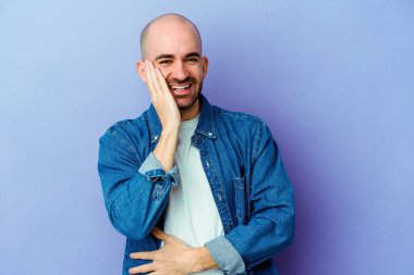 Mor arka planda izole genç beyaz kel adam mutlu bir şekilde gülüyor ve ellerini karnının üstünde tutarak eğleniyor..