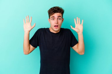 Kafkasyalı genç adam mavi arka planda izole edilmiş gökyüzüne haykırıyor, yukarı bakıyor, hayal kırıklığına uğramış..