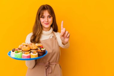 Sarı arka planda izole edilmiş genç pasta şefi kadın parmağıyla bir numarayı gösteriyor..