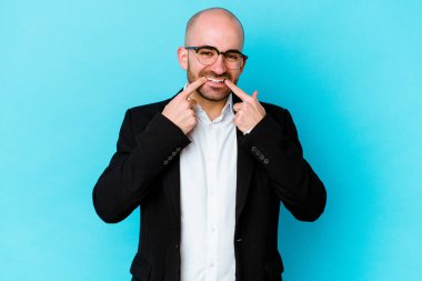 Genç, beyaz, kel bir adam mavi arka planda izole edilmiş gülümsüyor ve parmaklarını ağzına doğrultuyor..
