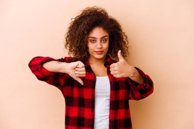 Genç Afrikalı Amerikalı kadın bej arka planda izole edilmiş. Baş parmağını kaldırıyor ve baş parmaklarını eğiyor. Zor seçim konsepti.