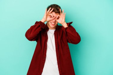 Mavi arka planda izole edilmiş genç beyaz adam bir başarı fırsatı bulmak için gözlerini açık tutuyor..