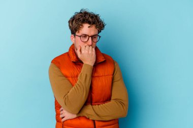 Mavi arka planda izole edilmiş, tırnak yiyen, gergin ve endişeli gözlüklü genç bir beyaz adam..