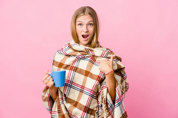 年轻的俄罗斯女人裹在毯子里喝咖啡 手指着咖啡 面带微笑 惊讶极了 — 图库照片