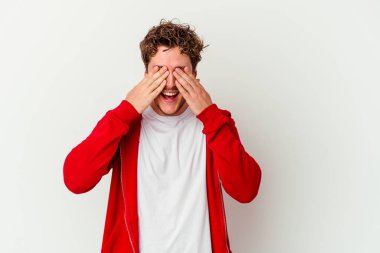 Beyaz arka planda soyutlanmış beyaz tenli genç bir adam elleriyle gözlerini kapatıyor, geniş bir gülümsemeyle bir sürpriz bekliyor..