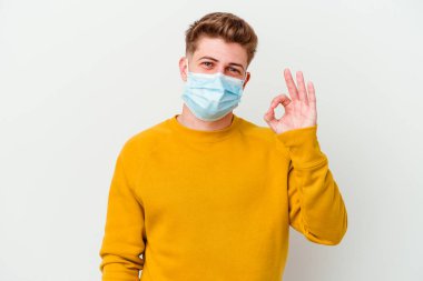 Koronavirüs maskesi takan genç adam beyaz arka planda izole edilmiş neşeli ve kendinden emin bir şekilde iyi niyet gösteriyor..