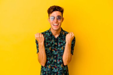 Hawaii gömleği giyen genç beyaz adam sarı arka planda izole edilmiş tasasız ve heyecanlı bir şekilde tezahürat yapıyor. Zafer konsepti.