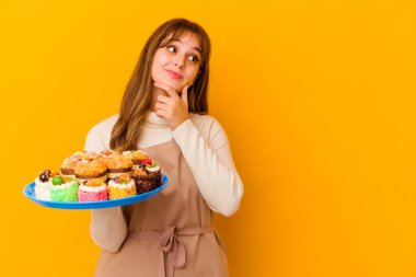 Sarı arka planda izole edilmiş genç pasta şefi kadın şüpheli ve şüpheci bir ifadeyle yana bakıyor..