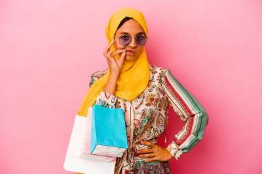 Genç Müslüman kadın pembe arka planda izole giysiler alıyor. Parmakları dudaklarında sır saklıyor..