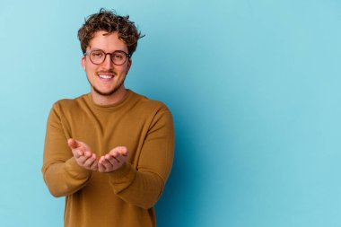 Mavi arka planda izole edilmiş gözlük takan genç, beyaz bir adam dudaklarını katlıyor ve hava öpücüğü yollamak için avuç içleri tutuyor..