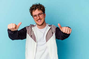 Mavi arka planda gözlüklü genç beyaz adam iki parmağını da kaldırıyor, gülümsüyor ve kendinden emin..