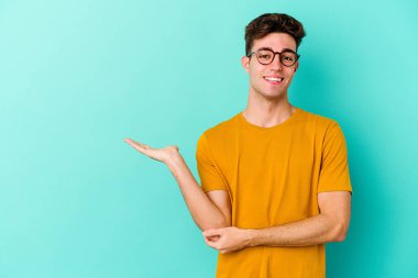 Mavi arka planda izole edilmiş genç beyaz bir adam bir palmiye üzerinde fotokopi alanı ve belinde bir el daha gösteriyor..