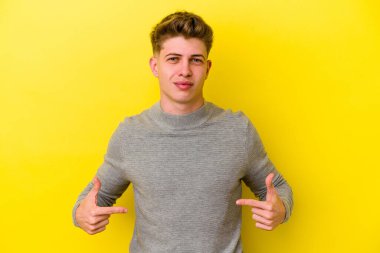 Sarı arka planda izole edilmiş genç beyaz bir adam gömlek fotokopi alanını elle gösteriyor, gururlu ve kendinden emin.