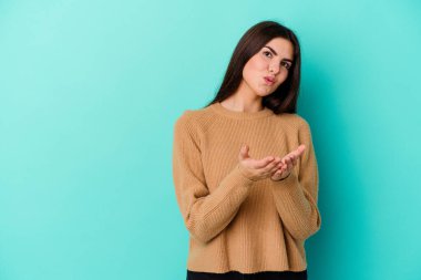 Mavi arka planda izole edilmiş genç, beyaz bir kadın. Dudaklarını kıvırıyor ve avuç içlerine hava öpücüğü yolluyor..