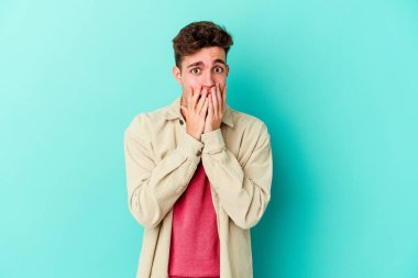 Mavi arka planda izole edilmiş genç beyaz adam şok olmuş, ağzı ellerle kaplanmış, yeni bir şey keşfetmeye hevesli..