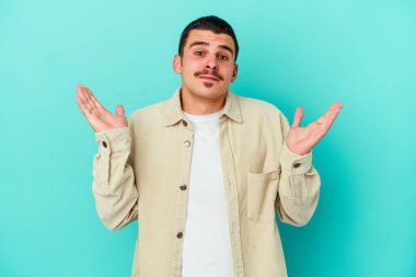 Kafkasyalı genç adam mavi arka planda izole edilmiş. Kafası karışmış ve omuz silkiyor. Kopya alanını tutmak için..