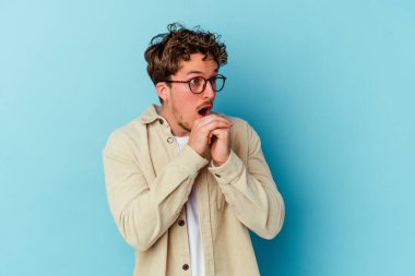 Mavi arka planda izole edilmiş gözlüklü genç beyaz adam şans için dua ediyor, şaşırıyor ve ağzını açıyor..