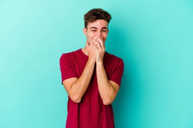 Mavi arka planda izole edilmiş genç beyaz bir adam bir şeye gülüyor, ağızlarını ellerle kapatıyor..