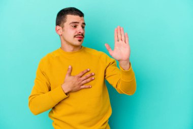 Kafkasyalı genç adam mavi arka planda izole edilmiş yemin ediyor, elini göğsüne koyuyor..