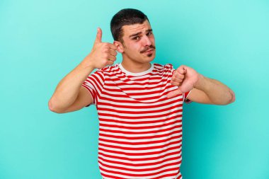 Kafkasyalı genç adam mavi arka planda izole edilmiş, baş parmağını kaldırıyor, zor seçim konsepti.