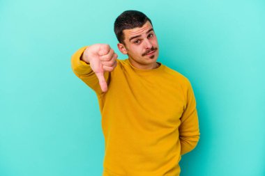 Mavi arka planda izole genç beyaz adam baş parmağını aşağı gösteriyor, hayal kırıklığı konsepti.