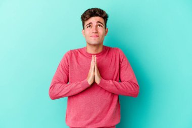 Mavi arka planda izole edilmiş genç beyaz bir adam el ele tutuşup, ağzının kenarında dua ediyor, kendinden emin..