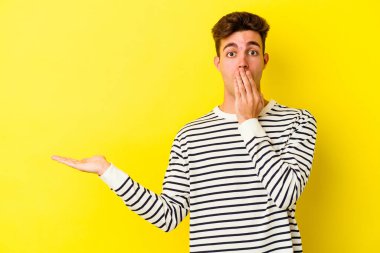 Sarı arka planda izole edilmiş genç beyaz adam, avuç içi fotokopi alanını elinde tutmaktan etkilendi..