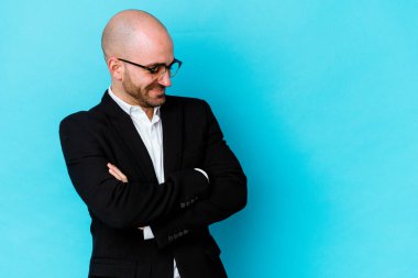 Genç, beyaz, kel bir adam mavi arka planda izole edilmiş, kollarını kavuşturarak gülümsüyor..