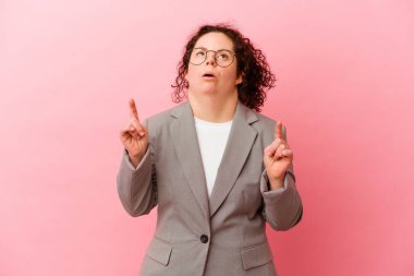 Down sendromlu bir iş kadını pembe arka planda izole edilmiş ve ağzı açık bir şekilde üst tarafı işaret ediyor..