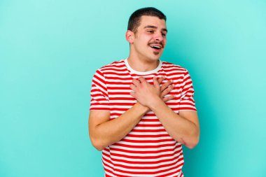 Kafkasyalı genç adam mavi arka planda izole olmuş gülüyor, elini yüreğine koyuyor, mutluluk kavramını koruyor..