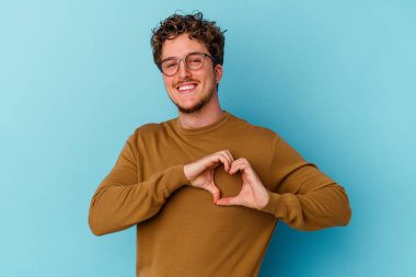 Mavi arka planda izole edilmiş gözlük takan genç beyaz adam gülümsüyor ve elleriyle kalp şeklini gösteriyor..