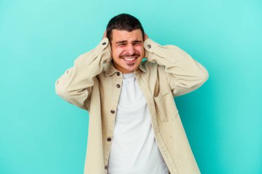 Mavi arka planda izole edilmiş genç beyaz adam kulaklarını elleriyle kapatıyor..