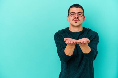 Kafkasyalı genç adam mavi arka planda izole edilmiş dudaklar kıvrılıyor ve hava öpücüğü göndermek için avuç içleri tutuyor..
