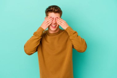 Mavi arka planda soyutlanmış genç beyaz bir adam elleriyle gözlerini kapatıyor, geniş bir gülümsemeyle bir sürpriz bekliyor..