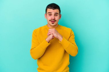 Kafkasyalı genç adam mavi arka planda izole edilmiş şans için dua ediyor, şaşırmış ve ağzını açıp önüne bakıyor..